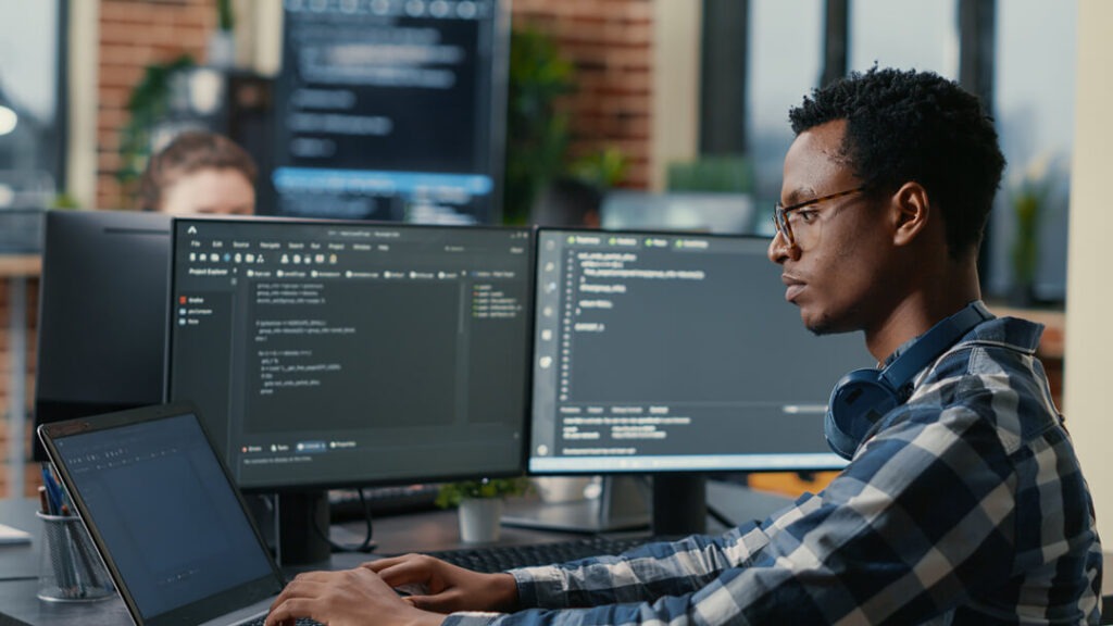 Desenvolvedor negro trabalhando em um computador, com outras duas telas ao lado, ajudando no gerenciamento de TI.
